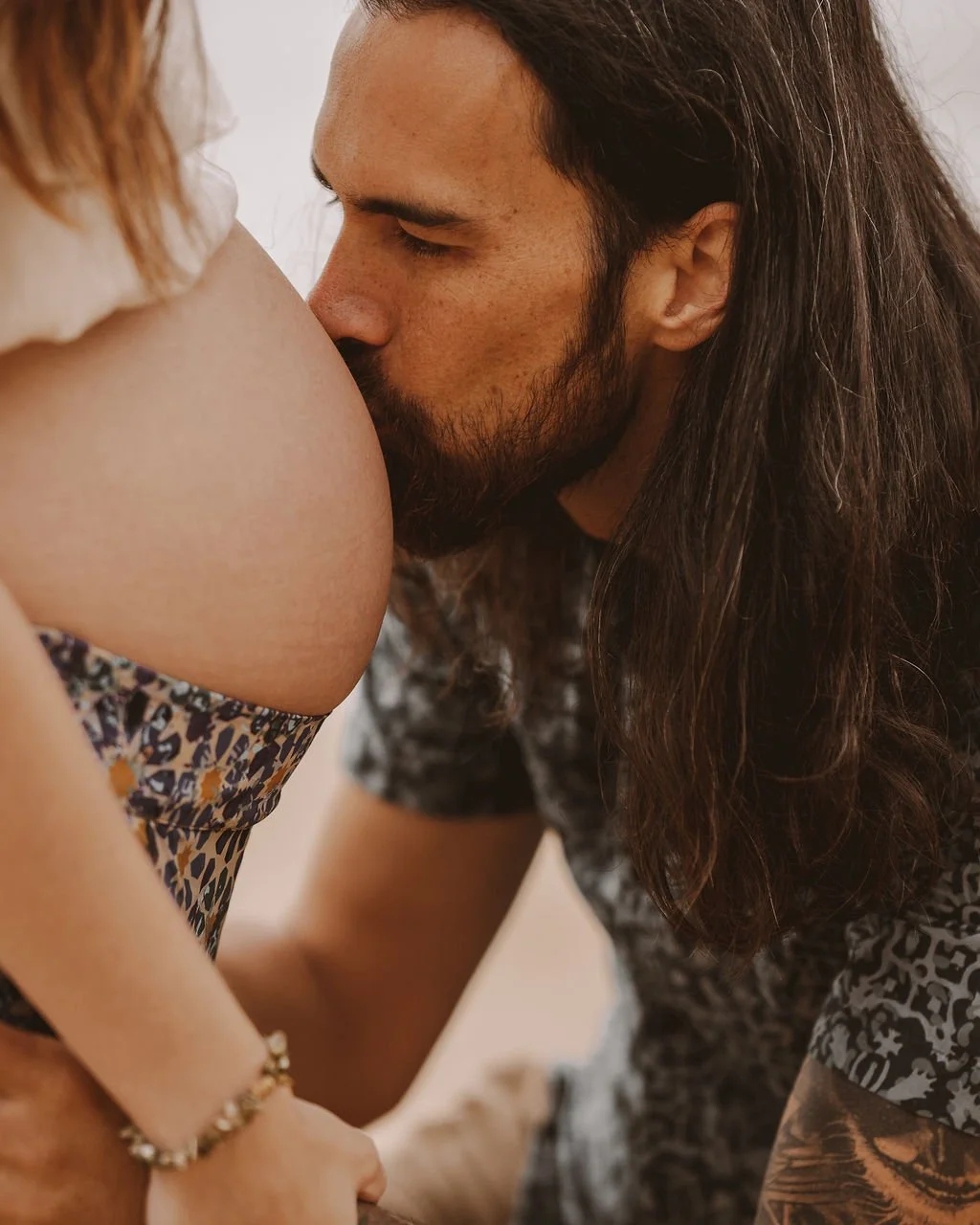 A man with long dark hair and a beard kisses a pregnant woman's belly.