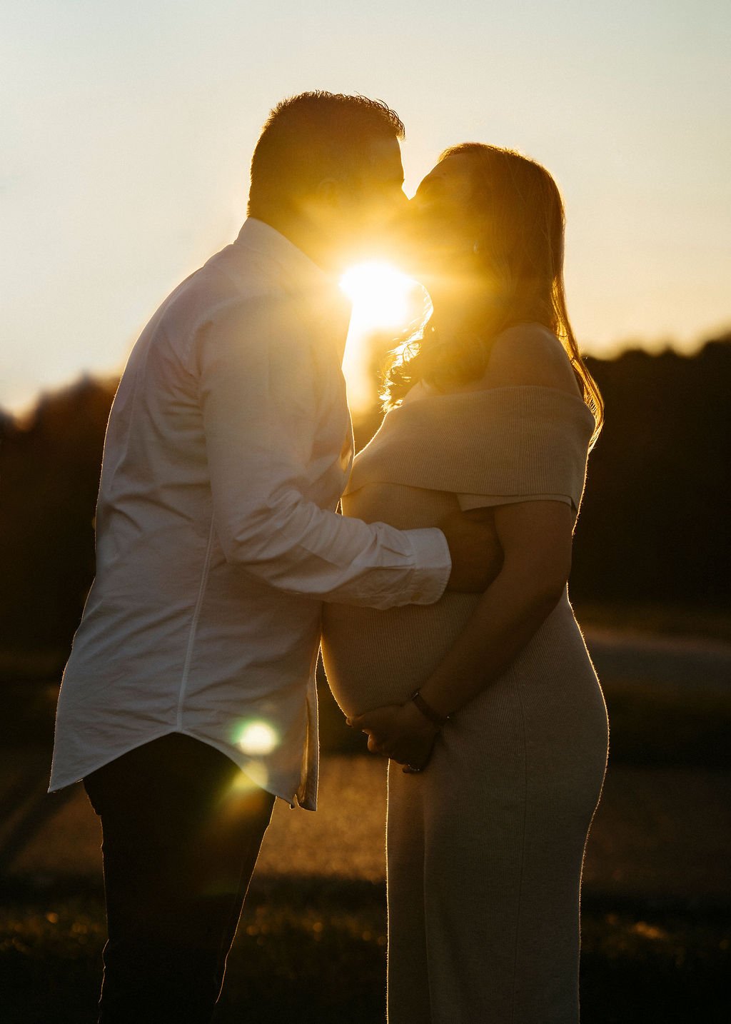 A couple kissing at sunset, with the pregnant woman's belly visible, outdoors during the golden hour.