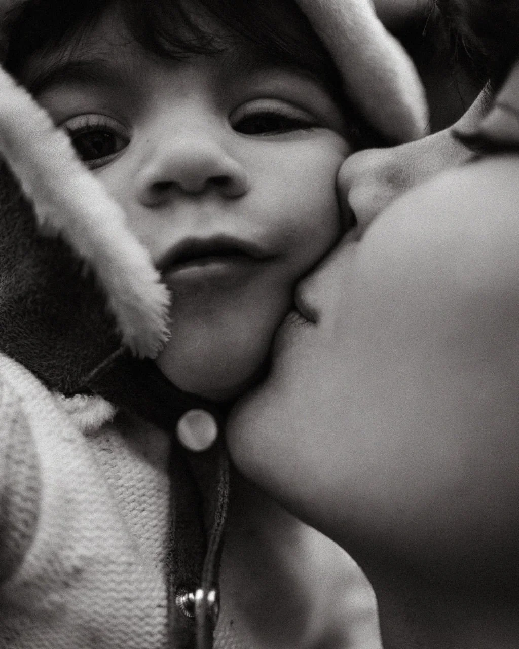 Close-up black and white photo of a young child with someone kissing her on the cheek.