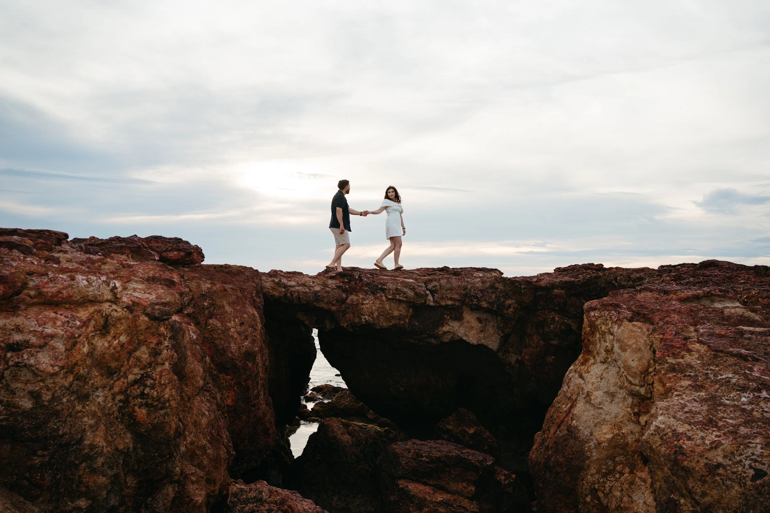 A couple holding hands on a large rock formation, with the ocean below and an overcast sky overhead.