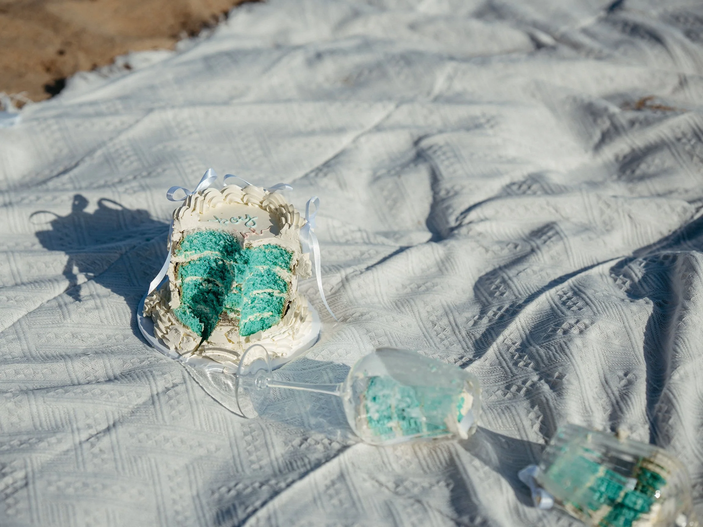 A partially eaten round cake with white frosting and blue-green filling inside, sitting on a white textured cloth outdoors, with a small decorative object nearby.