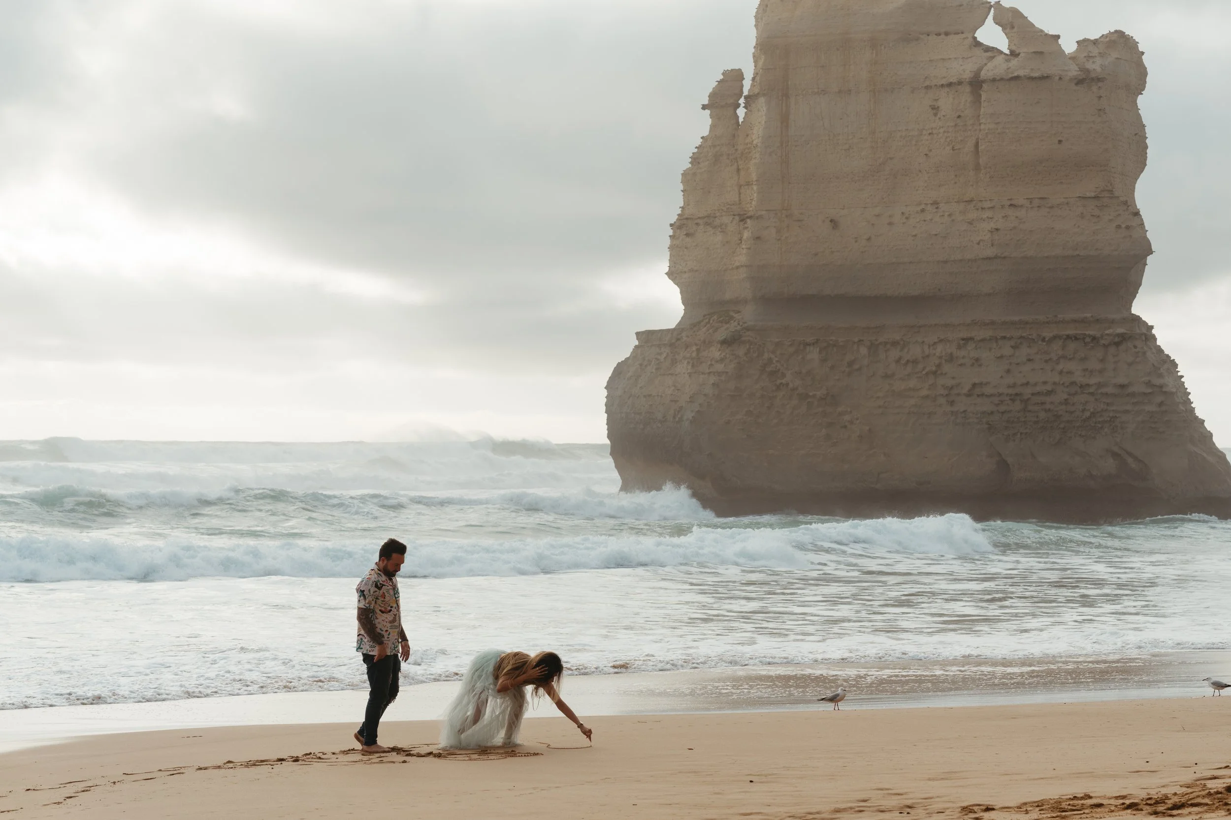 A man, woman, and child are on a beach in front of water with large rock formations and cliffs in the background. The woman, dressed in a white dress, is bending down to draw or write in the sand. The man stands nearby, watching her. The sky is overcast.
