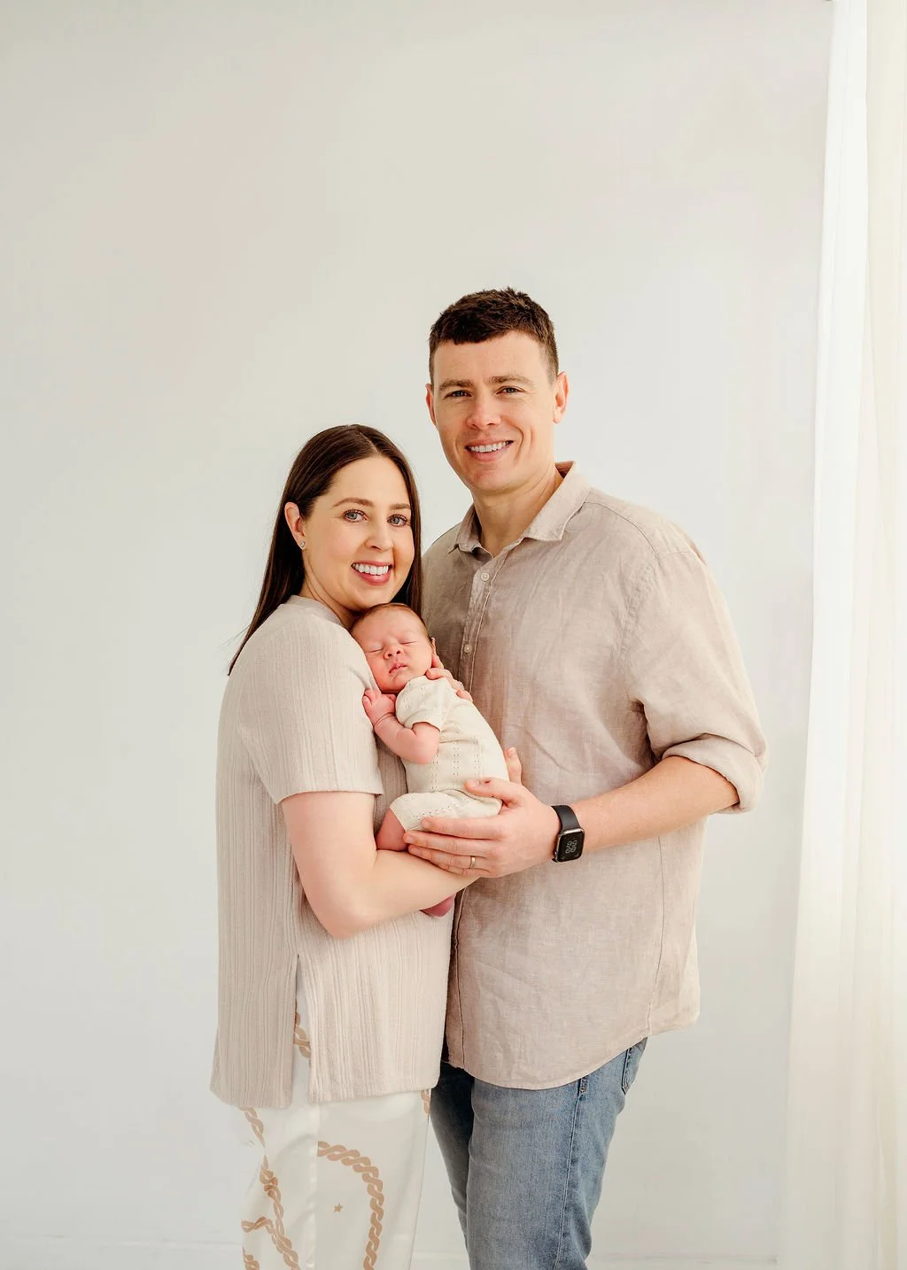 A happy family of three with a newborn, standing indoors against a plain light background, smiling at the camera.