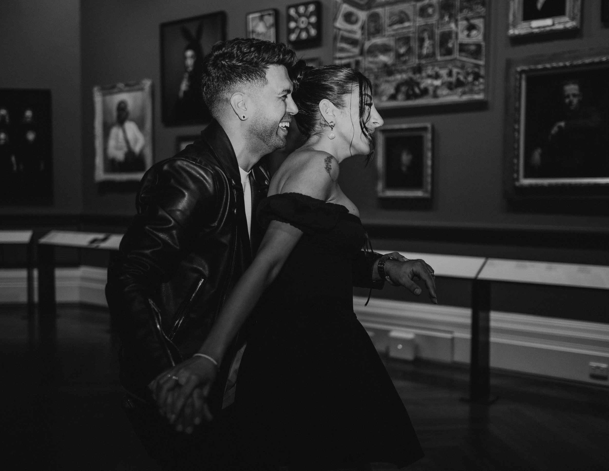 A black and white photo of a man and woman sharing a joyful moment, possibly laughing, with an art gallery wall in the background.