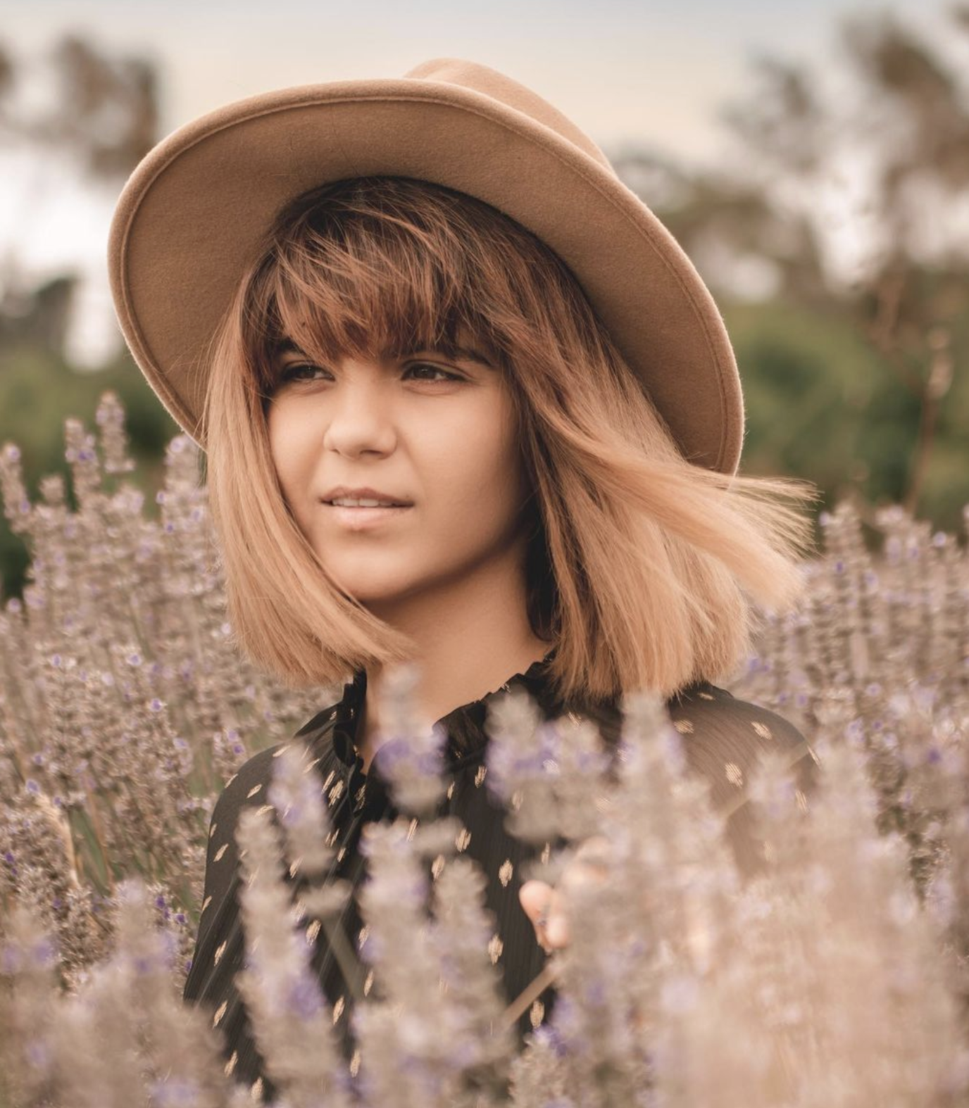 A woman in a tan wide-brimmed hat and black dress stands among lavender flowers in a field, tilting her head slightly to the side.