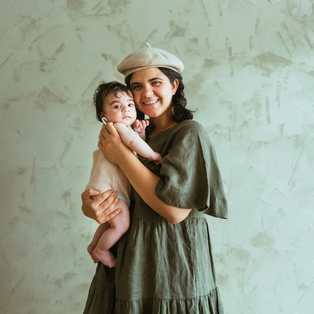 A woman smiling while holding a baby, both looking at the camera, standing in front of a textured light green wall. The woman is wearing a beige beret and a loose olive green dress. The baby has dark hair and is wearing a light-colored sleeveless outfit.