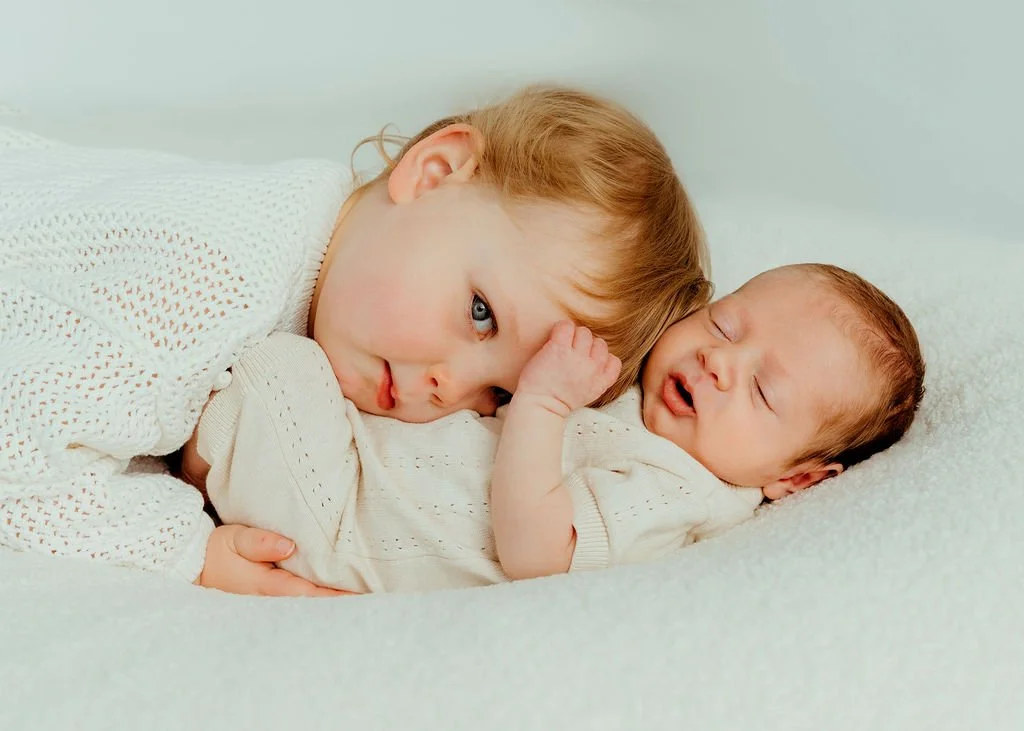 A young girl with red hair lying on a soft surface, resting her head beside a sleeping newborn baby. The girl has her arm around the baby and looks softly at the baby.
