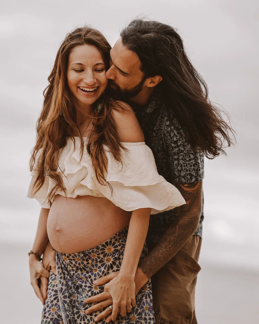 A pregnant woman with long red hair smiling as a man with long dark hair kisses her on the cheek, both standing outdoors against a cloudy sky.