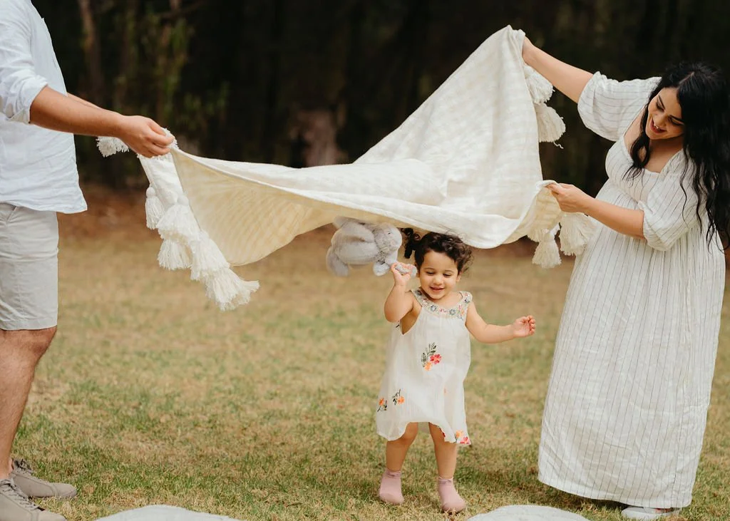 A family of three playing with a blanket outdoors on grass, with the little girl holding a stuffed animal and smiling.