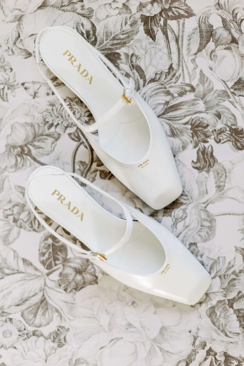Pair of white Prada high-heeled shoes with ankle straps on a floral-patterned surface.