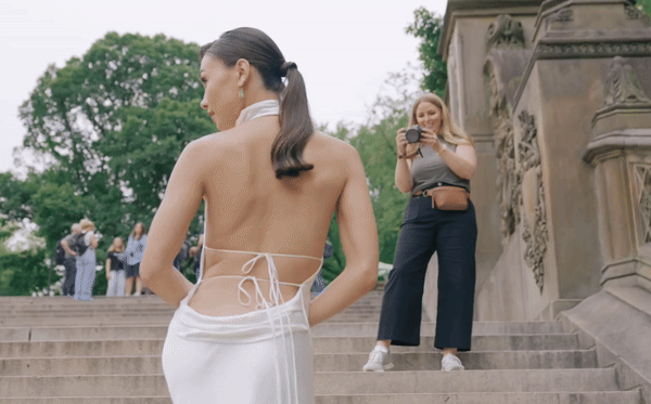 A woman in a white dress is posing for photos on outdoor steps, while another woman with a camera takes her picture. There are trees and other people in the background.