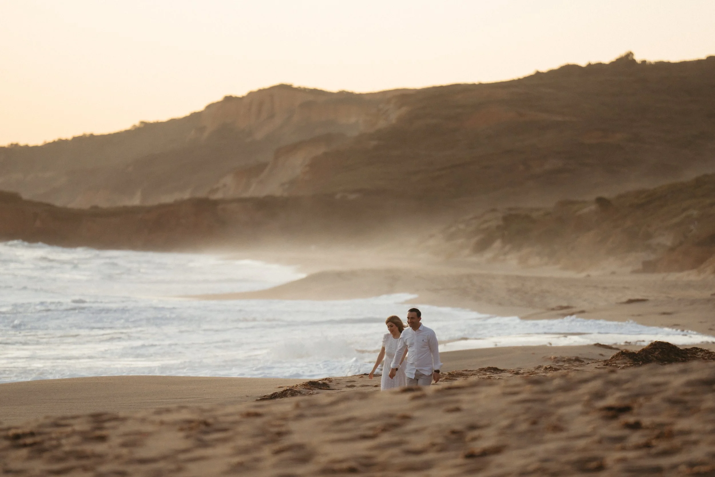 A couple walking along the beach during sunset, with rocky cliffs in the background.