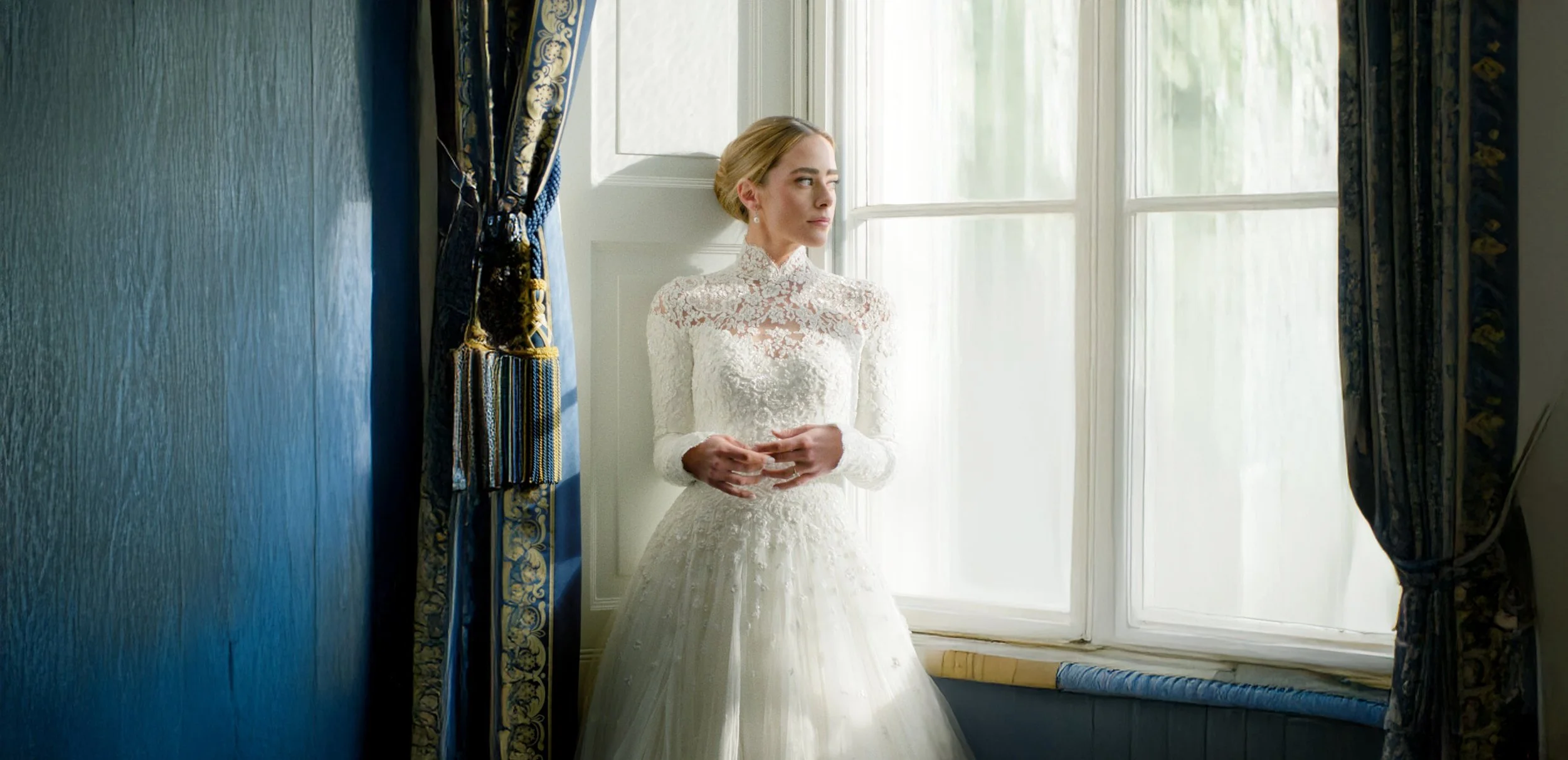 A young woman in a vintage white lace wedding dress stands by a window, holding her hands together, looking contemplatively outside.