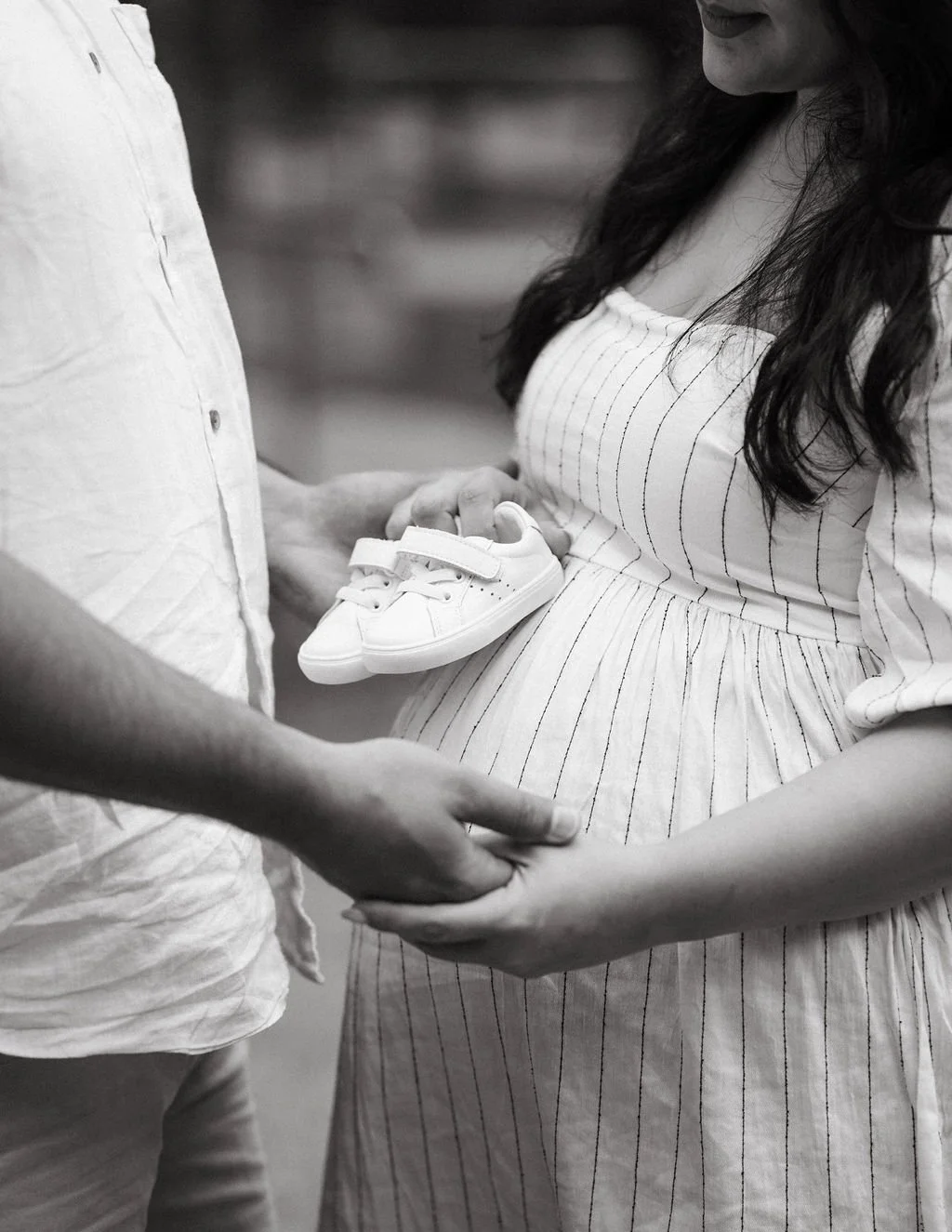 Pregnant woman in a light-colored, striped dress holding tiny baby shoes, with partner's hand touching her pregnant belly.