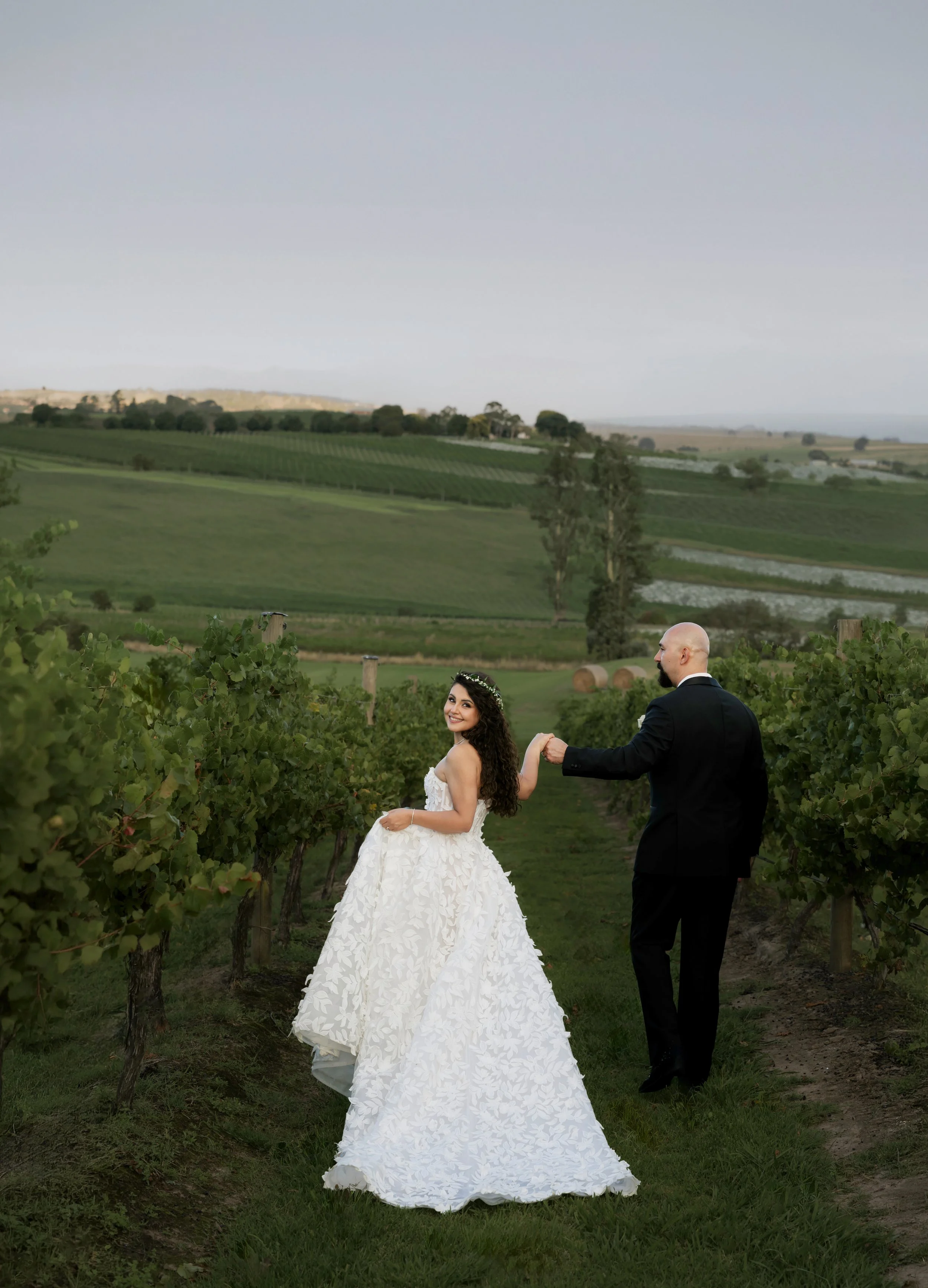 A newlywed couple holding hands and smiling outdoors. The groom is in a black tuxedo with a bow tie, and the bride is in a white wedding dress holding a bouquet of flowers. There are trees and a gazebo in the background.