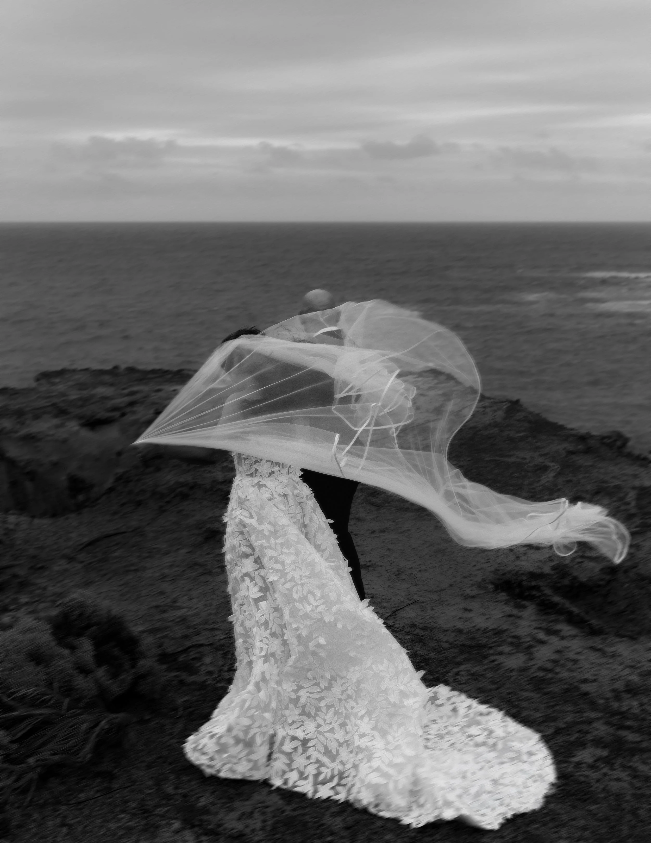 Black and white photo of a woman in a lace dress holding a transparent, flowing umbrella on a rocky beach, with ocean and cloudy sky in the background.
