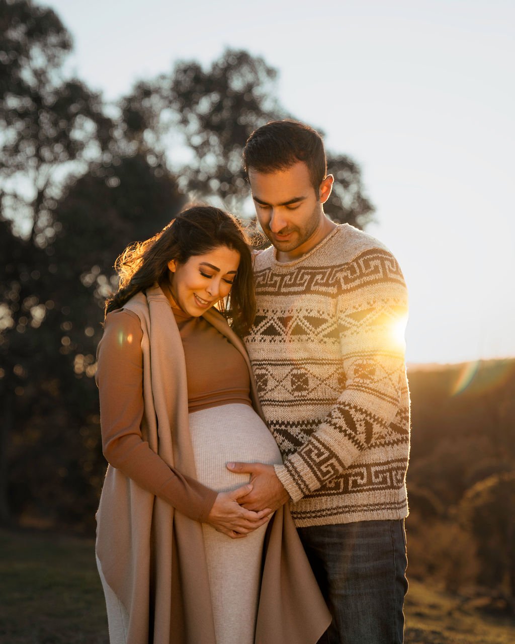 A pregnant woman and a man standing outdoors at sunset, both smiling and touching her belly.