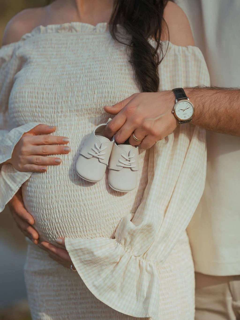 A pregnant woman wearing a yellow checkered off-shoulder dress, holding a pair of baby shoes in front of her belly. A man, wearing a wristwatch and a wedding ring, is placing the shoes in her hands. The woman's hand rests on her belly, and part of hi