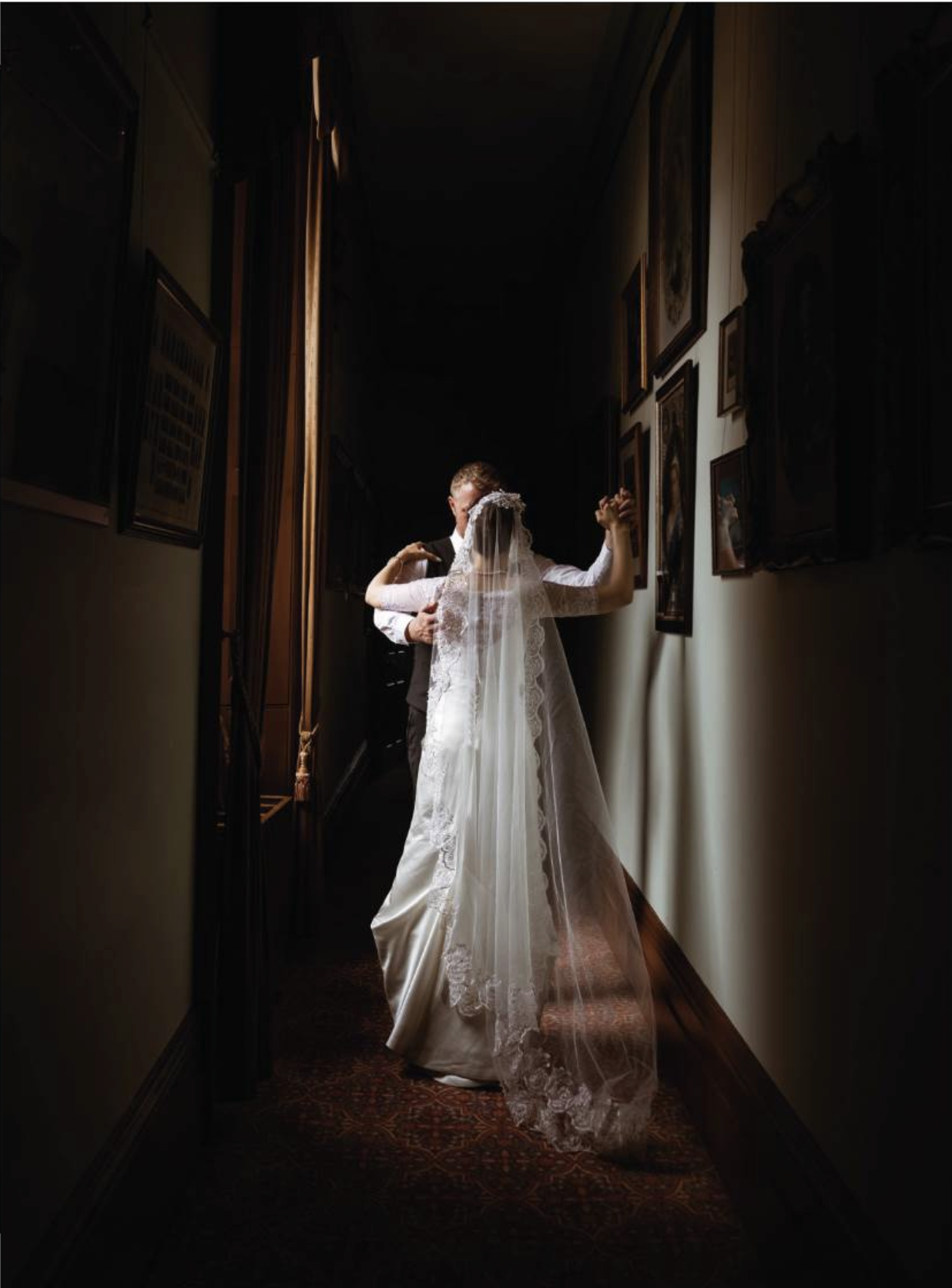A bride in a wedding dress and veil standing in a dimly lit hallway, facing away, with a groom behind her, holding her hand.