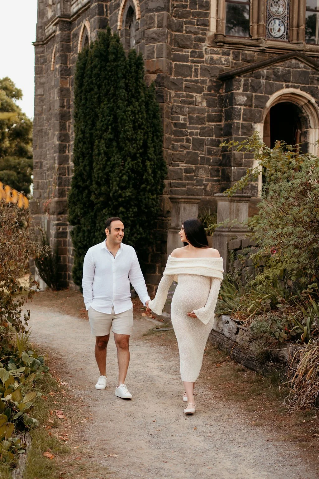 A couple walking hand in hand on a dirt path outside an old stone church, with lush plants and trees around them.