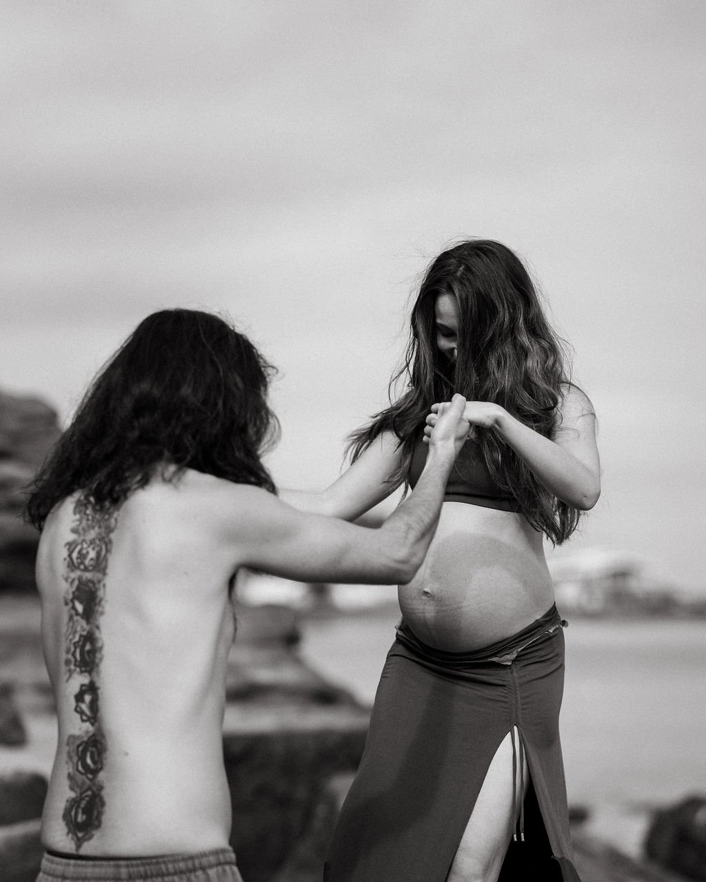 A woman with long hair, appearing pregnant, standing on a beach and holding hands with a man with long hair who has tattoos on his back.