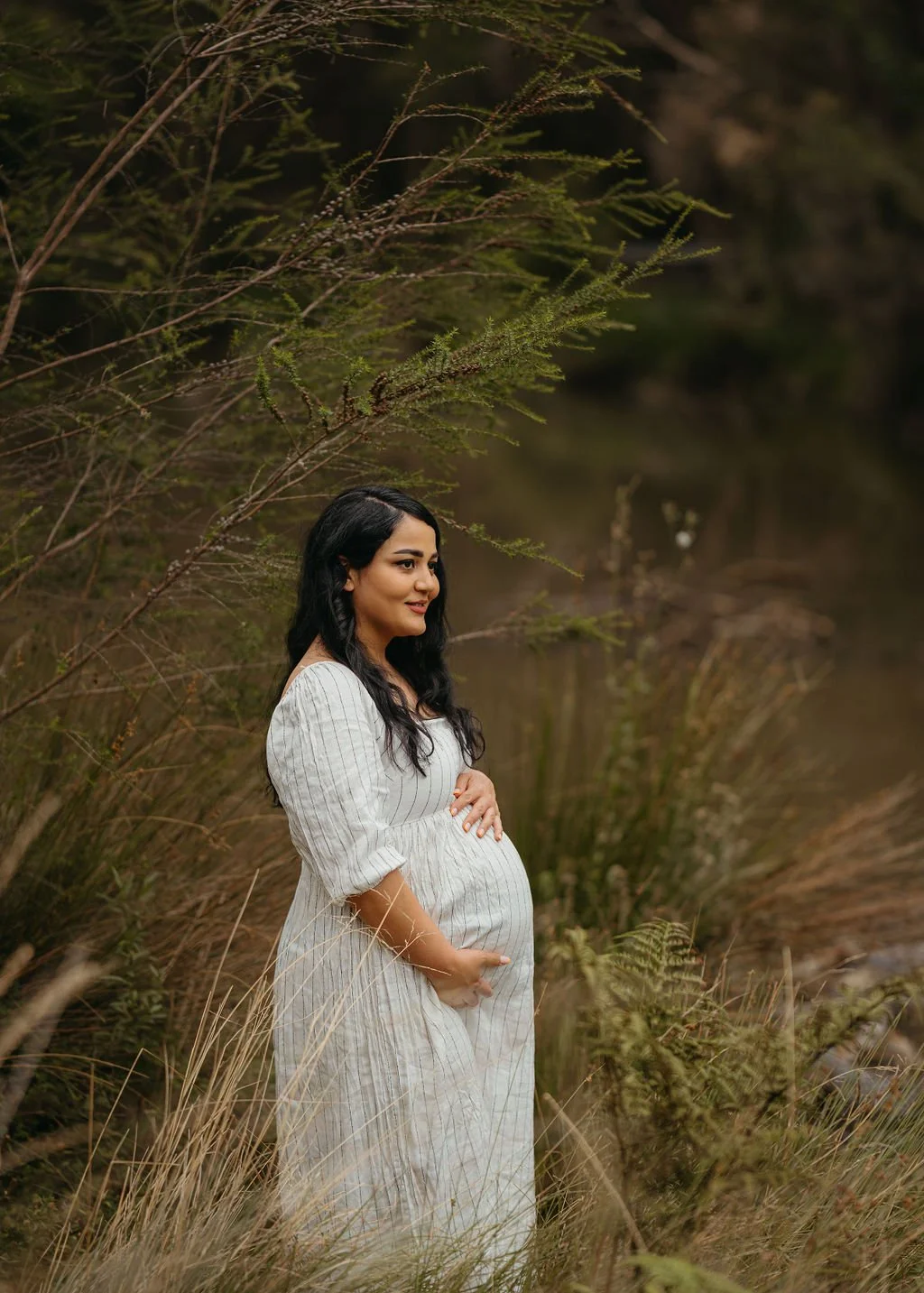 A pregnant woman with long black hair in a white, striped dress standing outdoors among tall grasses and trees, gently holding her belly with a peaceful expression.