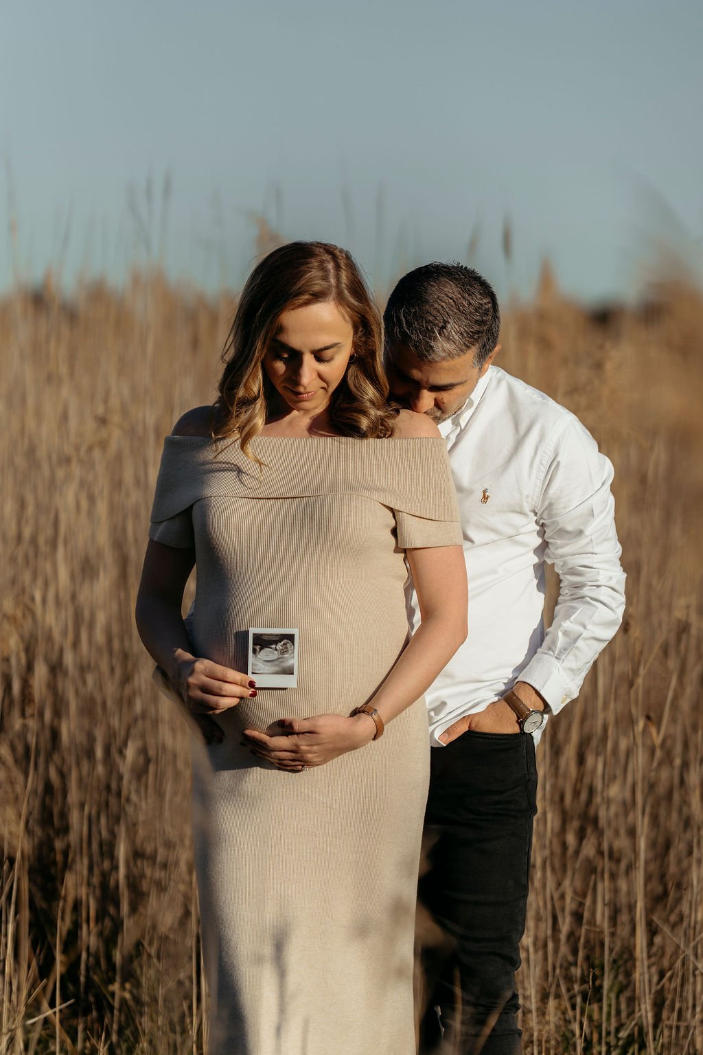 A pregnant woman holding an ultrasound photo, standing in a field with a man behind her, both looking down, during sunset.
