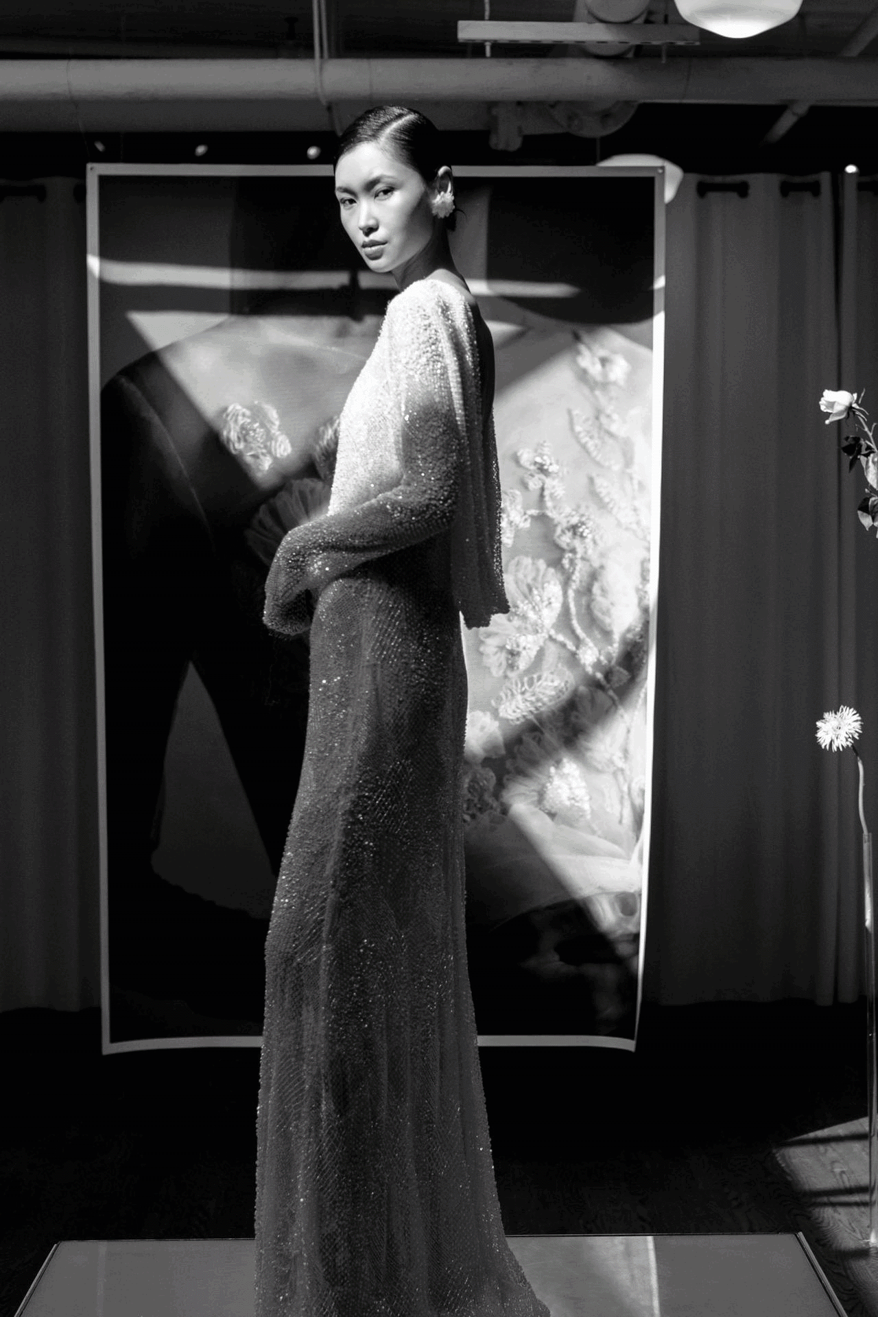 A woman in a shimmering, sheer gown stands in front of a large floral backdrop, with a single flower on a stand to her right, in a black-and-white photo.
