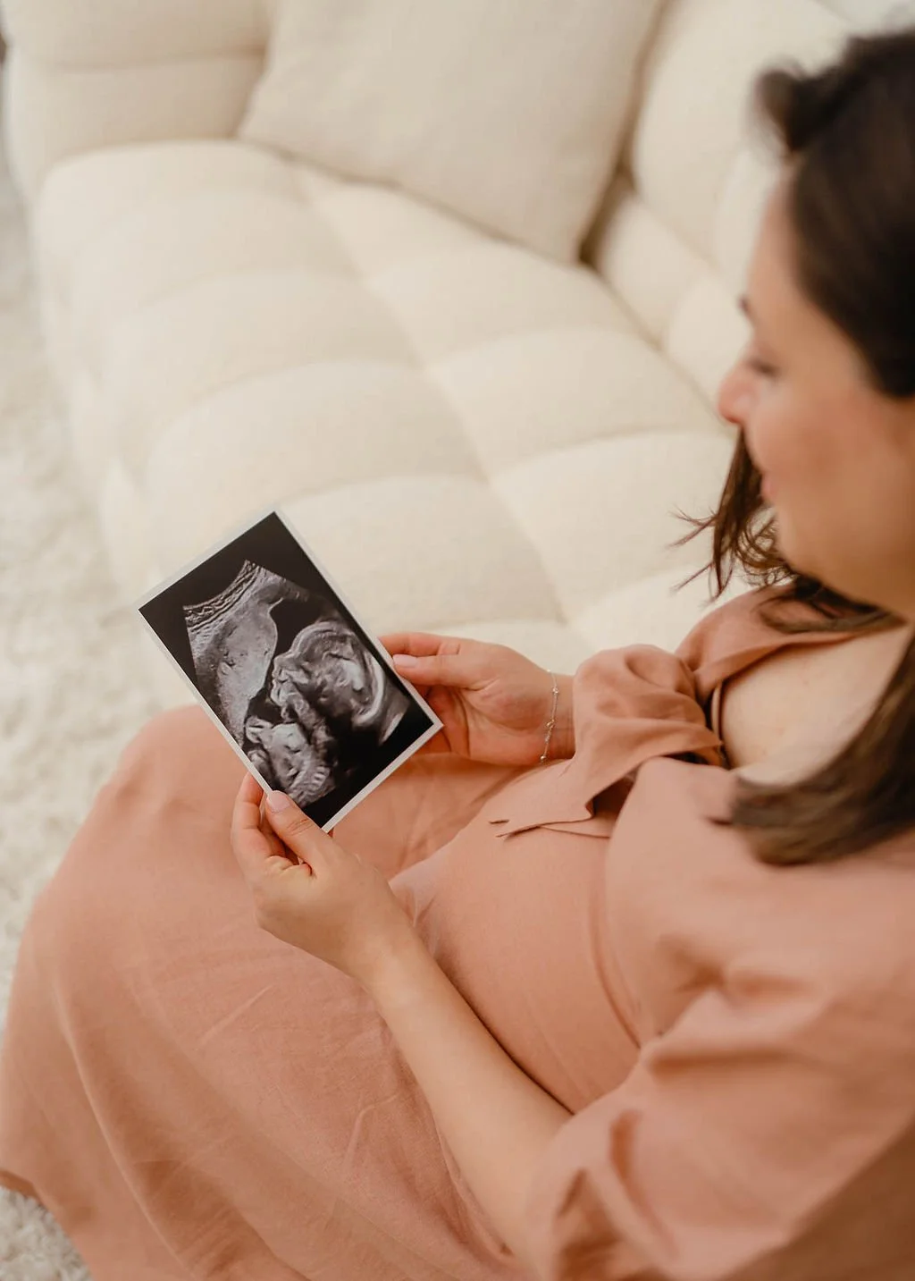 Woman sitting on a beige couch holding an ultrasound photo of a developing baby.