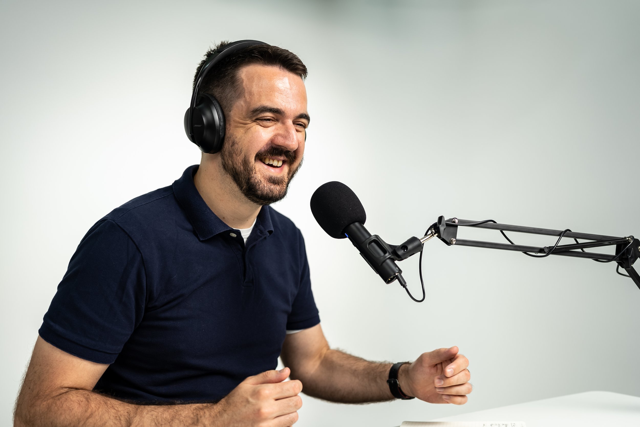 A man with dark hair and a beard, wearing headphones, speaking into a microphone, smiling, and sitting at a table with a white background.