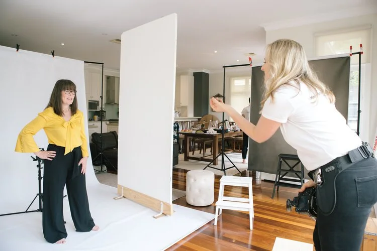 Two women in a photography studio setup; one is posing and another is taking a photograph.