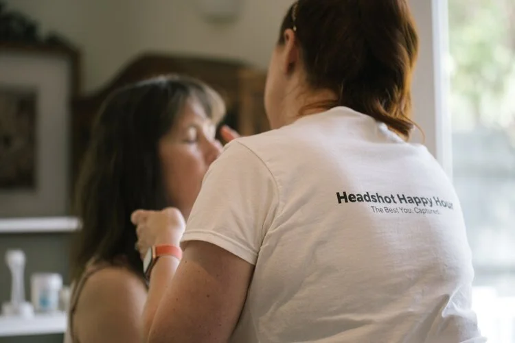 A makeup artist applying makeup to a woman's face, wearing a T-shirt that says "Headshot Happy Hour" in a well-lit room.