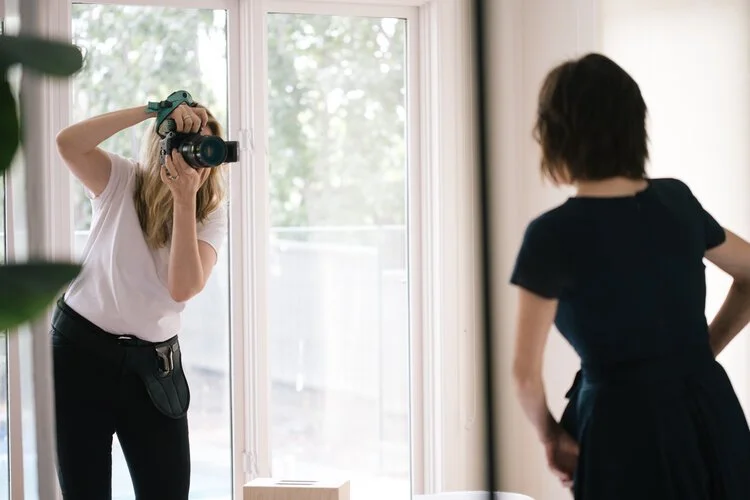 Photographer taking a picture of a person indoors near a large window, with natural light shining through.
