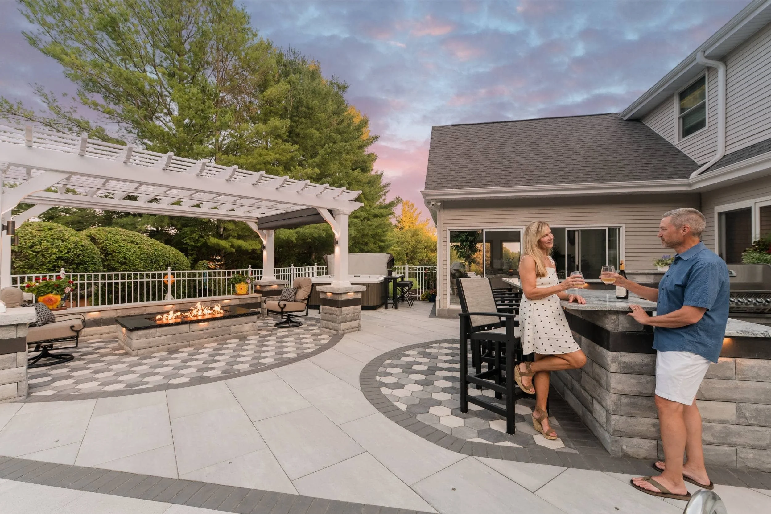 Mand and woman drink wine at outdoor kitchen bar on paver patio with lit fire pit nearby