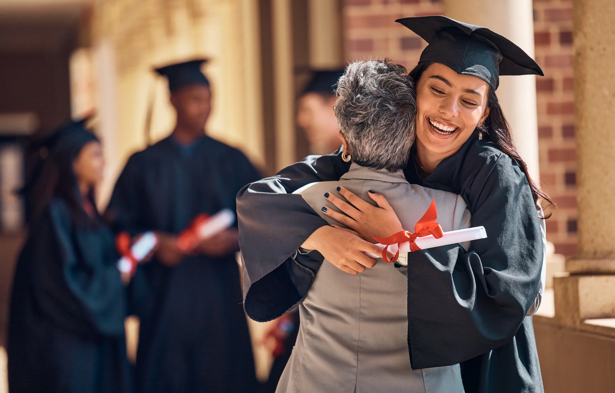Graduate in cap and gown hugging an older woman on graduation day with other graduates in the background