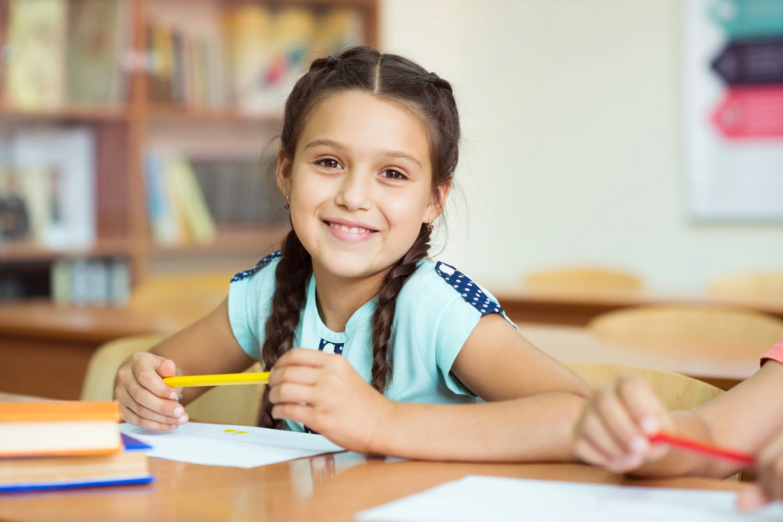 A smiling young girl with braided hair sitting at a desk in a classroom, holding a yellow pencil, with notebooks and papers in front of her.