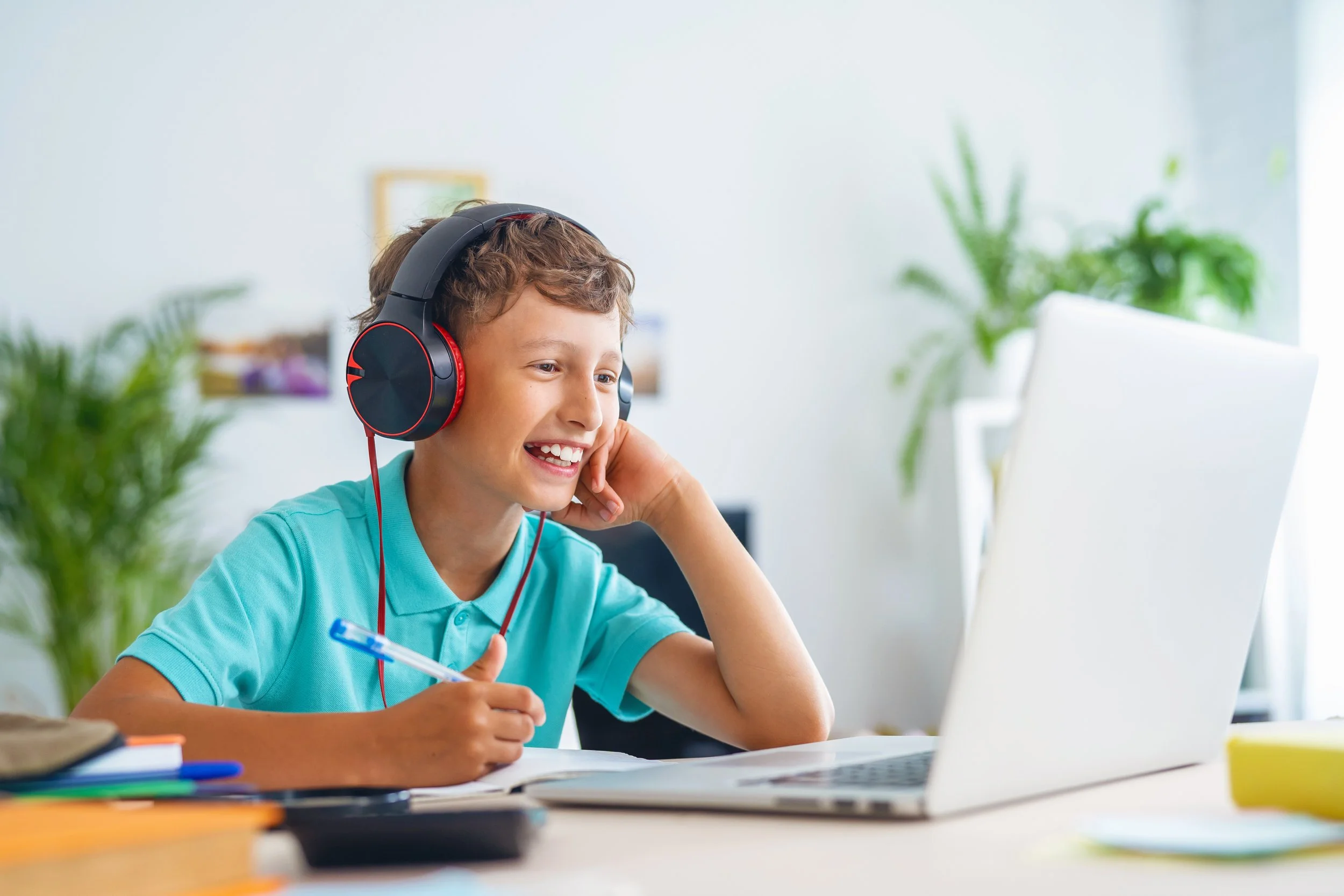 A young boy with brown hair wearing red and black headphones, smiling while looking at a laptop screen, with a pen in his hand, sitting at a desk with books and notebooks, in a brightly lit room with green plants.