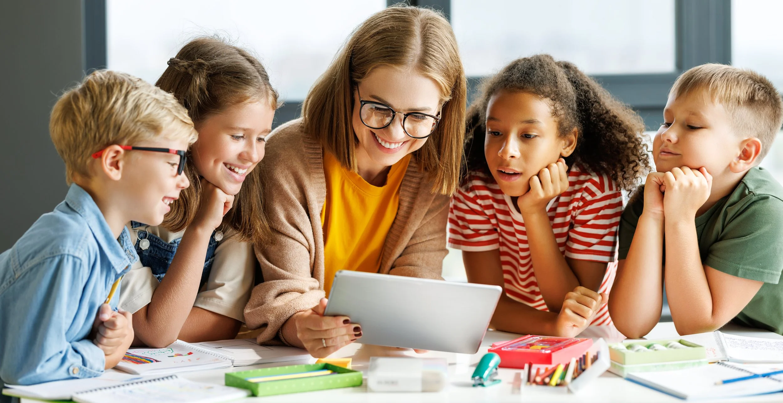A smiling woman with glasses shows a tablet to a group of five children seated at a table with school supplies, engaging in a learning activity.