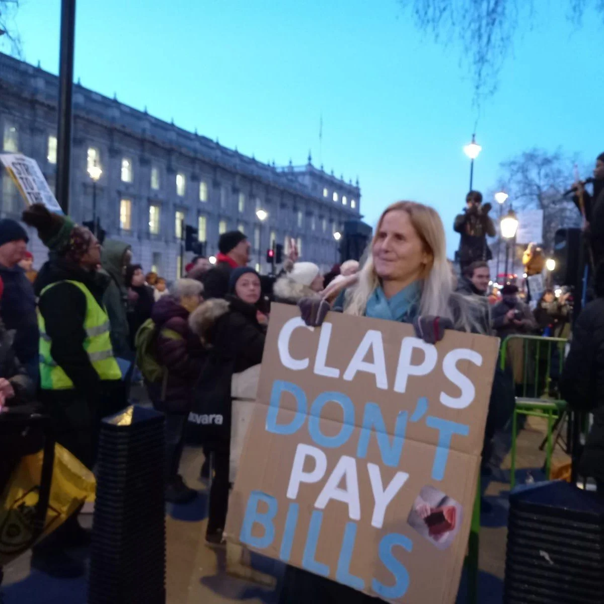 Woman holding a protest sign that reads 'Claps Don't Pay Bills,' amidst a crowd of protesters in an urban setting during evening.