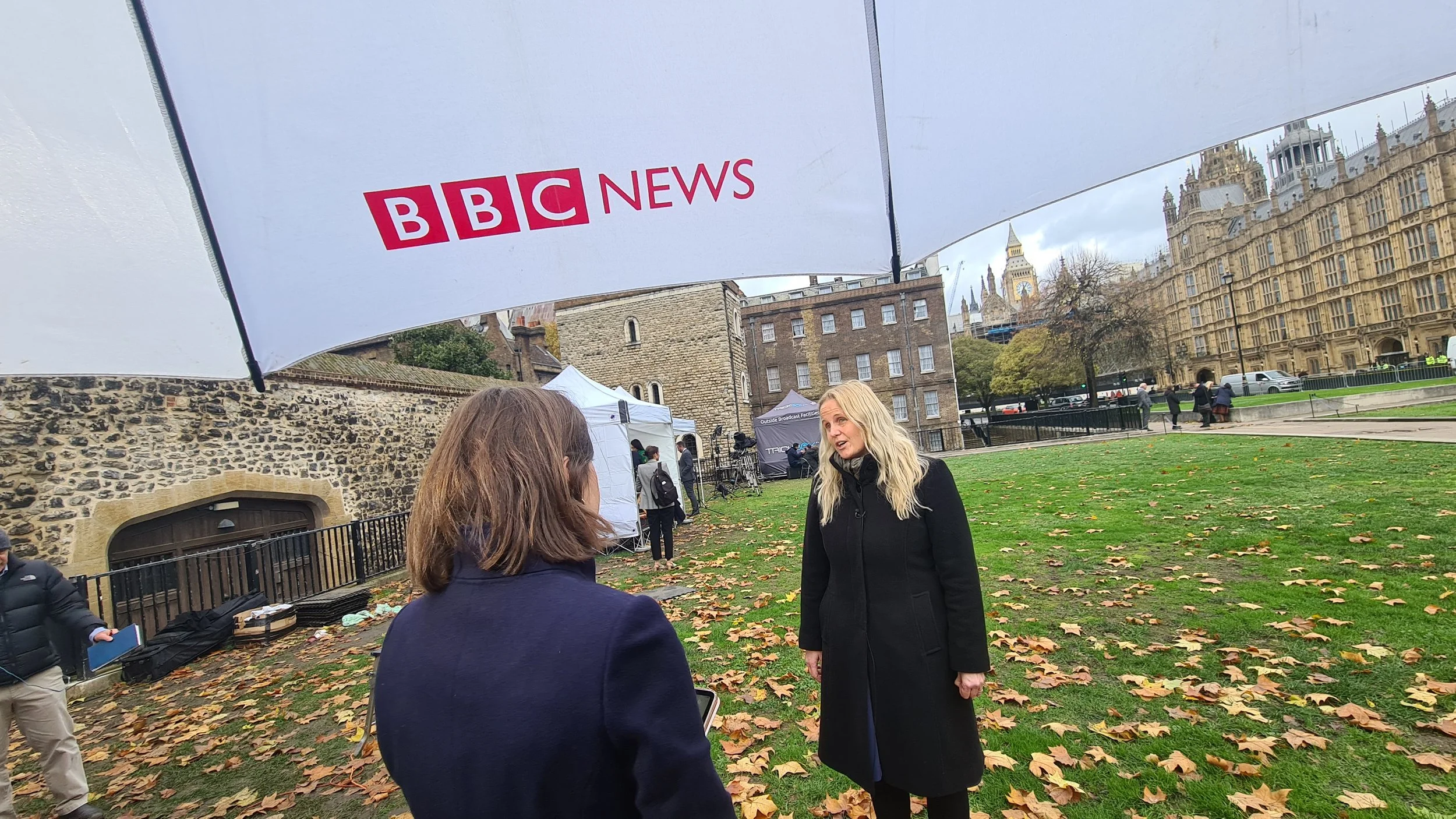 Two women talking outdoors under a BBC News umbrella with fallen autumn leaves on the ground and historical buildings in the background.