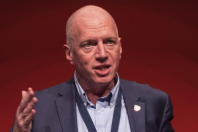 A bald man wearing a blue suit and shirt, speaking on stage with a red background.