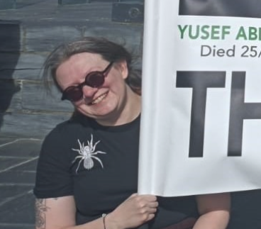 Woman smiling, wearing sunglasses and a black t-shirt with a spider graphic, holding a sign.