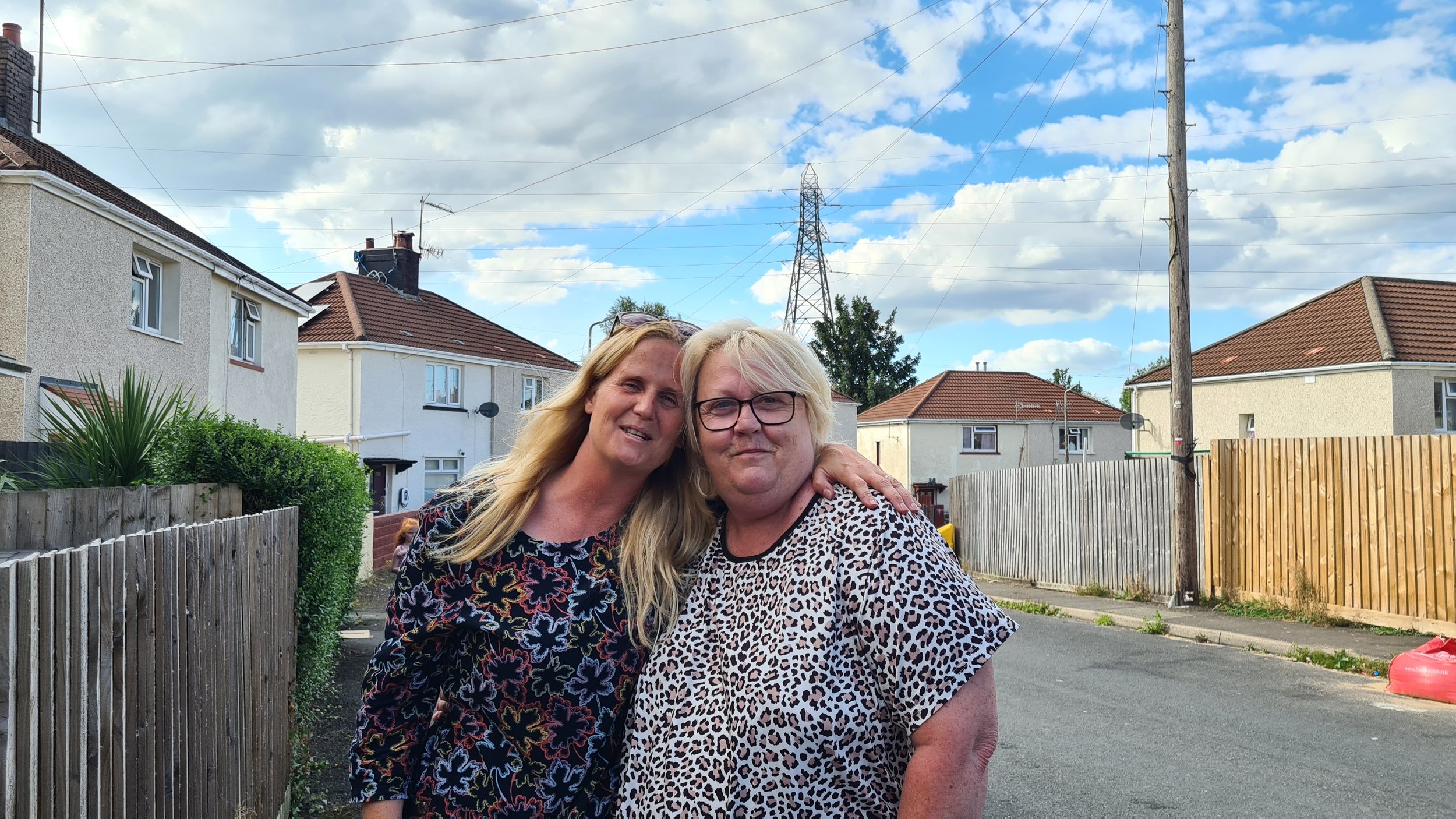 Two women standing close together on a residential street, smiling and embracing, with houses and a cloudy sky in the background.