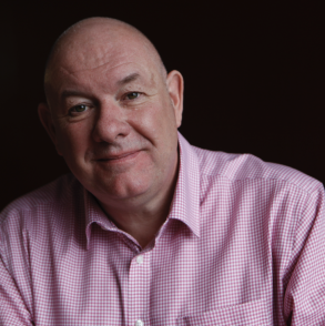 A middle-aged man with a friendly expression, bald head, wearing a pink and white checkered shirt, against a dark background.