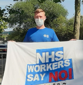 A man wearing a face mask holding a banner that reads 'NHS WORKERS SAY NO!' in front of trees on a sunny day.