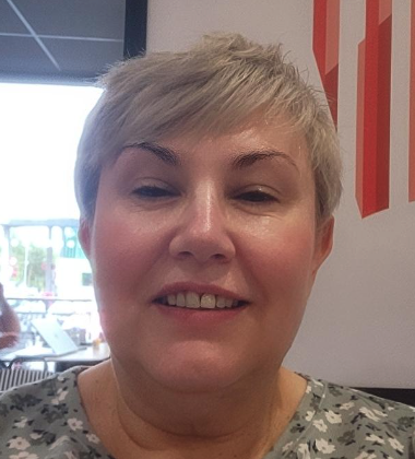 A woman with short blonde hair and a patterned top smiling indoors near a window and a red and white wall.