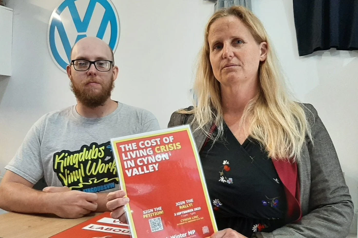 Two people sitting at a table, holding a red poster that reads 'The Cost of Living Crisis in Cynon Valley, Join the Rally, 3rd September 2022, 11am - 1pm, Labour' with a Volkswagen logo in the background.