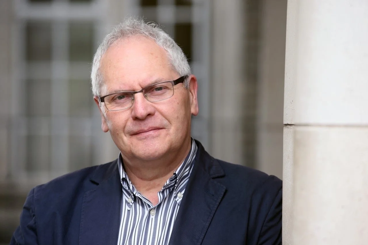 A middle-aged man with gray hair, wearing glasses, a striped shirt, and a dark blazer, standing outdoors near a stone building.