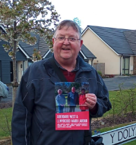 A smiling man holding a behind-the-scenes pass badge in front of houses in a neighborhood.