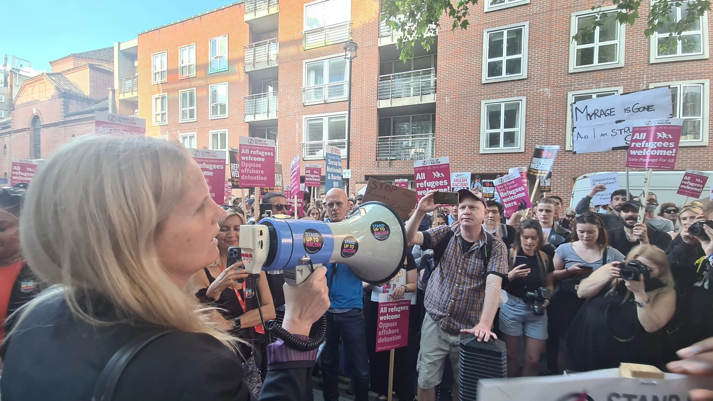 Crowd of protesters holding signs and banners, some with messages like "All refugees welcome!" and "Stop racism." A woman in the foreground is speaking into a megaphone. In the background, there are residential buildings and trees.